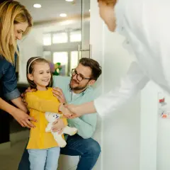 Uśmiechnięta dziewczynka z misiem wita się z ortodontką, kiedy z dzieckiem do ortodonty. Rodzice wspierają ją.