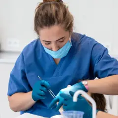 Dentystka w niebieskim fartuchu i masce zakłada opatrunek na zęba pacjentowi.