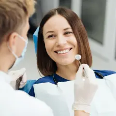 Uśmiechnięta pacjentka podczas wizyty u dentysty. Lekarz w masce i rękawiczkach bada jej zęby, przygotowując się do ewentualnego piłowania zębów.