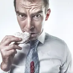 Mężczyzna w białej koszuli i krawacie je pączka, ubrudzony cukrem pudrem. Zastanawia się, ile nie jeść po plombie, gdy dżem ląduje na jego krawacie.