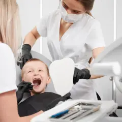 Uśmiechnięty chłopiec u dentysty, który bada jego wędzidełko języka. Asystentka trzyma lusterko w kształcie zęba.