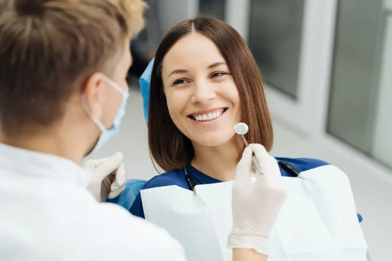Uśmiechnięta pacjentka podczas wizyty u dentysty. Lekarz w masce i rękawiczkach bada jej zęby, przygotowując się do ewentualnego piłowania zębów.