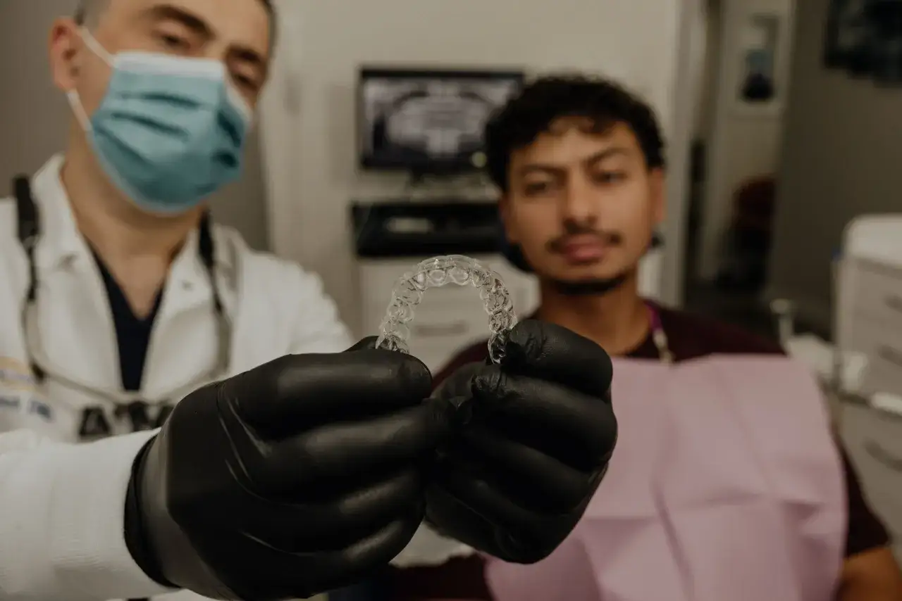 Dentysta w czarnych rękawiczkach pokazuje pacjentowi przezroczyste nakładki. To jest właśnie Invisalign, co to jest?